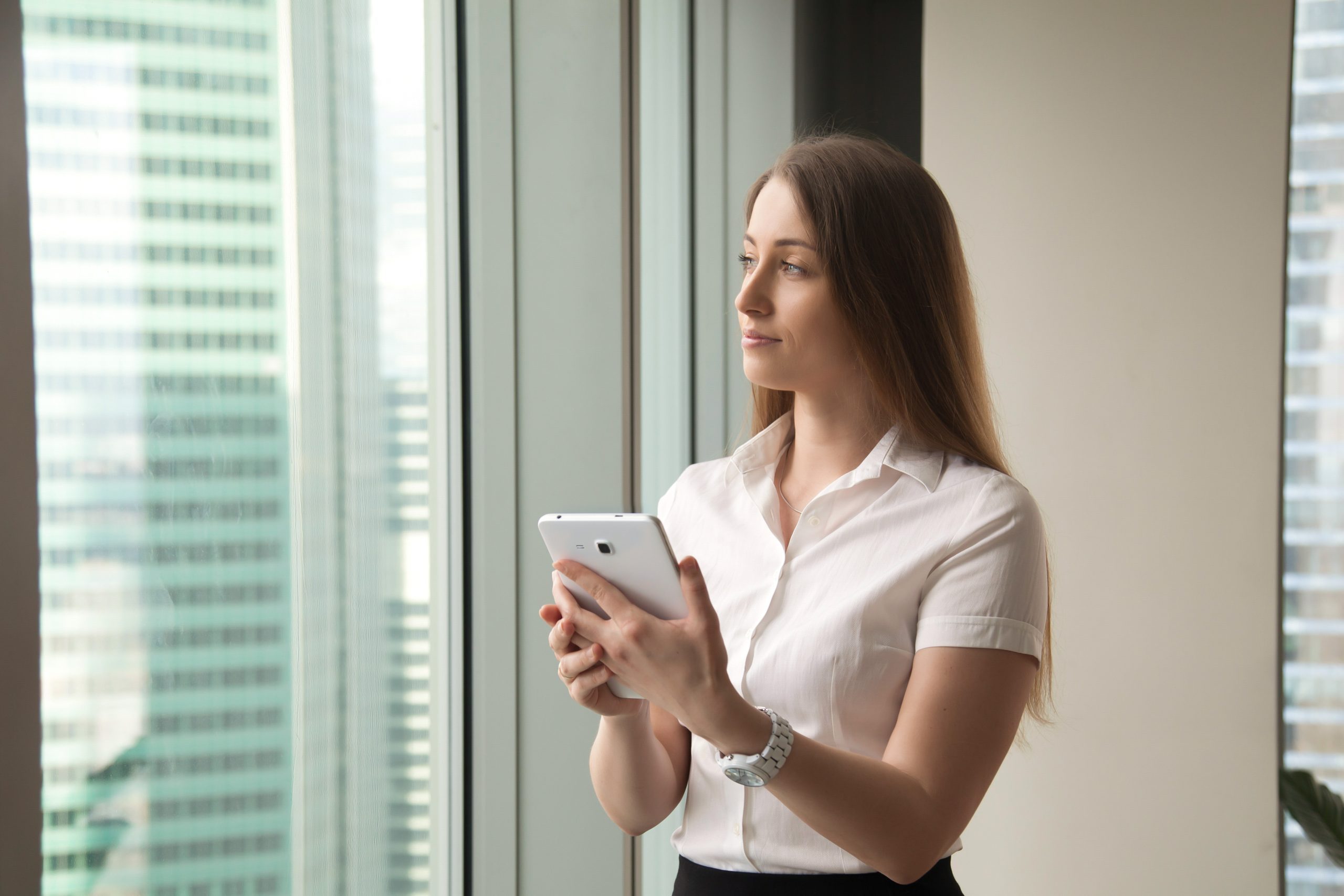 Young dreamy beautiful businesswoman looking through window whil